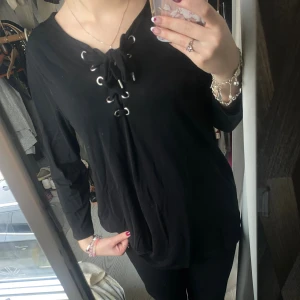 Svart långärmad topp med snörning från Vila - Svart långärmad topp från Vila med cool snörning och metallöljetter vid halsen. Toppen har en rundad halsringning och är gjord i mjuk bomull som känns skön mot huden. Perfekt för dig som gillar en enkel men edgy stil.