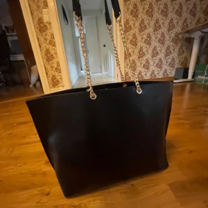 Svart väska  - En stilren snygg svart väska med kedjeremmar i guld. Det är några fläckar i väskan, se bild 2. Läderremarna som sitter på guldremmarna har något enstaka stygn gått upp på ( se bild 3) men funkar att använda. Inte toppkvalitet därav det låga priset:)        Bredd: 50 cm Höjd: 33cm