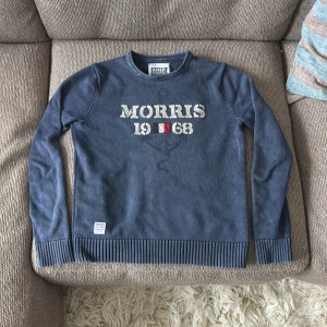 Blå tröja från Morris med tryck - Snygg blå tröja från Morris med broderat tryck 'Morris 1968' och flagga på bröstet. Tröjan har rund hals, ribbade muddar och är långärmad. Perfekt för dig som gillar klassisk och stilren design.