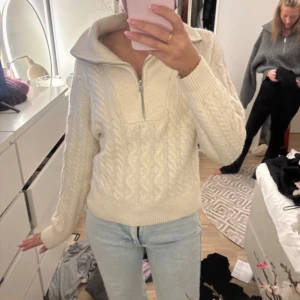 Stickad offwhite tröja med half zip - Supermysig stickad tröja i offwhite med kabelstickat mönster och halv dragkedja vid kragen. Tröjan har lång ärm, bred krage och ribbade muddar. Perfekt för kyliga dagar och passar till jeans eller mjukisbyxor.