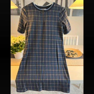 Mörkblå rutig klänning från edc-Esprit - Snygg mörkblå kortklänning från edc i rutigt mönster med rund hals och korta ärmar. Klänningen har en cool ringformad dragkedja framtill och ribbad kant vid halsen i blått. Perfekt för dig som gillar klassisk stil med en modern twist.