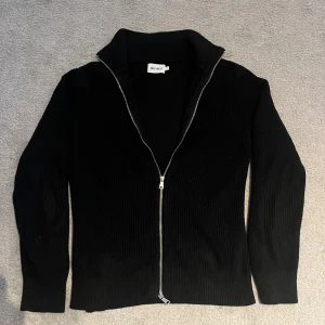 Svart kofta från East West - Riktigt fet svart ribbstickad kofta från East West med hel dragkedja framtill och krage. Tröjan har riktigt bra passform och är gjord i ett stickat material som känns både mjukt och bekvämt. Pris kan diskuteras.