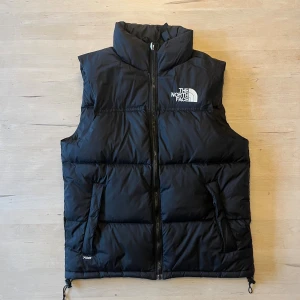 Svart dunväst från The North Face XS - Svart dunväst från The North Face med broderad logga på bröstet och ryggen. Västen har två sidofickor med dragkedja och justerbar nederkant med snörning. Perfekt för lager-på-lager. Storlek XS, men passar mig som brukar ha S. Väldigt sparsamt använd och mycket fint skick.