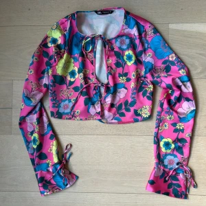 Blommig croppad blus från Zara - Säljer en färgstark croppad blus från Zara med blommigt mönster i rosa, blått, gult och grönt. Blusen har långa ärmar med knyt vid ärmslut och framtill, samt en snygg öppning vid halsen. Endast testad, nyskick