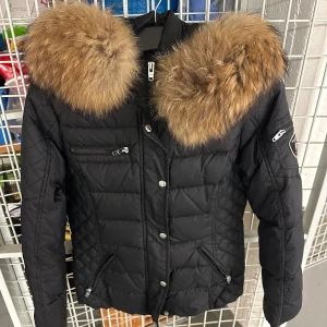 Rockandblue - Snygg svart pufferjacka med stor fluffig päls runt kragen. Jackan har flera dragkedjor och knappar framtill samt detaljer på ärmen. Perfekt för dig som vill ha en varm och trendig look under vintern.