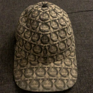 Beige keps från Salvatore Ferragamo - Snygg keps från Salvatore Ferragamo i beige och svart med ikoniskt mönster över hela kepsen. Tillverkad i bomull med detaljer i svart skinn baktill. Perfekt accessoar för att lyfta din outfit med en lyxig touch.