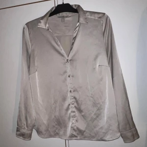 Champagnefärgad satinblus från H&M - Snygg champagnefärgad skjorta från H&M i glansigt satinliknande material. Klassisk krage, långa ärmar med manschett och knappar framtill. Perfekt för dig som gillar stilrena och trendiga plagg med en lyxig känsla.