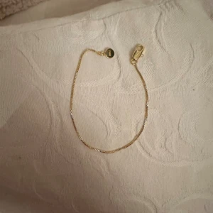 Guldarmband från Muli - Snyggt och tunt guldarmband från Muli med minimalistisk design. Armbandet har en rund bricka med Muli-loggan och en smidig karbinlås. Perfekt för dig som gillar stilrena och enkla smycken med guldfinish.