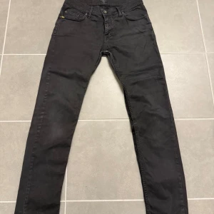 Svarta jeans från Tiger of Sweden - Svarta jeans från Tiger of Sweden med klassisk femficksdesign och smal passform. Jeansen har bälteshällor, knappgylf och diskret logga vid myntfickan. Perfekta för dig som gillar en stilren och enkel look. Storlek 29x32
