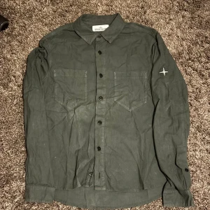 Mörkgrön skjorta från Stone Island - Snygg mörkgrön skjorta från Stone Island med klassisk krage och två bröstfickor. Skjortan har svarta knappar hela vägen och den ikoniska Stone Island-loggan broderad på vänster ärm. Tillverkad i mjuk bomull för skön passform.