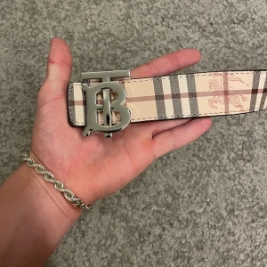 Burberry bälte med silverdetalj - Säljer ett brett Burberry-bälte i skinn med klassiskt rutigt mönster i beige, svart och rött. Bältet har en stor silvrig spänne med Burberrys ikoniska TB-logga. Ena sidan är beige med rutor och den andra helsvart med diskret rutstruktur och ingraverad logga.