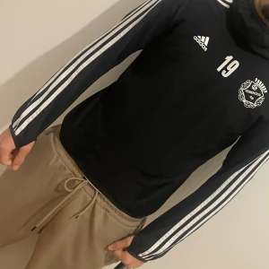 Svart Adidas träningströja med tryck - Svart långärmad träningströja från Adidas med vita ränder längs ärmarna och tryck på bröstet med nummer 19 och Veberöds logga. Hög krage och smal passform, perfekt för fotbollsträning eller sportiga dagar.