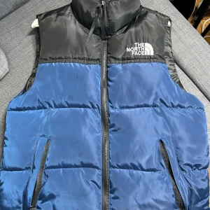 The North Face Dunväst - Snygg dunväst från The North Face i blått och svart med broderad logga på bröstet. Västen har hög krage, två sidofickor med dragkedja och quiltad design. Perfekt för lager-på-lager och outdoor vibes.