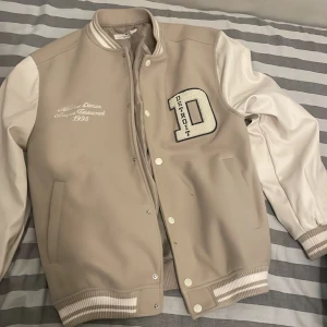 Beige och vit bomberjacka H&M - Snygg bomberjacka från H&M i beige med vita ärmar och ribbade muddar. Stor broderad D med texten Detroit på bröstet och broderad text på vänster sida. Jackan har knappar framtill och två fickor. Perfekt för dig som gillar college-stil.