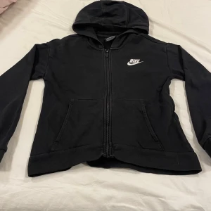 Svart Nike hoodie med dragkedja XL - Svart hoodie från Nike i storlek XL med vit logga på bröstet. Tröjan har huva, dragkedja framtill och två fickor. Tillverkad i mjuk bomullsblandning, perfekt för chill eller träning. Klassisk och enkel design som funkar året runt.