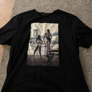 Svart Limitato t-shirt med print - Svart t-shirt från Limitato med stort konstnärligt print framtill som visar två personer vid ett träningsredskap med stadsmiljö i bakgrunden. T-shirten har rund hals och korta ärmar. Materialet är mjuk bomull och passformen är normal.