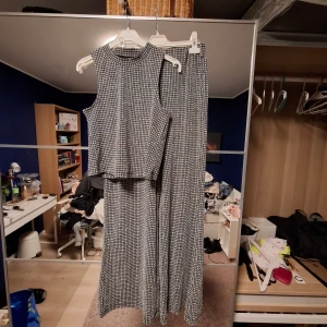 Fest set - Säljer ett par svarta och vita byxor med rutigt mönster och matchande crop top. Byxorna har en loose och wide passform med raka ben och elastisk midja och toppen är tight med polokrage. Materialet är mjukt och stretchigt, perfekt för en chill och trendig look. Passar dig som gillar att sticka ut med mönster. Byxorna i M och tröjan i S