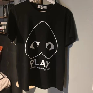 Cdg T-shirt - Svart t-shirt från Comme des Garçons Play med stort vitt tryck framtill som visar det ikoniska hjärtat med ögon och texten PLAY. Klassisk rund hals och korta ärmar. Materialet är mjuk bomull som känns skön mot huden.