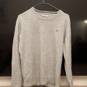 Ljusgrå tröja från GANT  - Säljer en stilren ljusgrå tröja från GANT med rund halsringning och diskret broderad logga på bröstet. Tröjan är långärmad och tillverkad i mjuk bomull, perfekt för en clean och klassisk look.