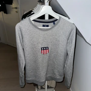 Grå sweatshirt från GANT med broderi - Snygg grå sweatshirt från GANT i bomull med broderad logga och emblem framtill. Tröjan har rund hals, långärmade raglanärmar och ribbade muddar. Perfekt för chill dagar och enkel att matcha med jeans eller joggers.