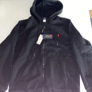 Svart hoodie från Polo Ralph Lauren - Svart hoodie från Polo Ralph Lauren med dragkedja och huva. Klassisk röd logga broderad på bröstet, snörning i huvan och två fickor framtill. Perfekt för chill dagar och enkel att matcha med jeans eller joggers.