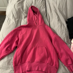 Rosa hoodie med huva och ficka - Säljer en chill rosa hoodie med stor magficka och huva. Tröjan har ribbade muddar vid ärmslut och nederkant, perfekt för att mysa eller hänga med kompisar. Materialet är mjukt och skönt, troligen bomull eller polyester. Passar dig som gillar färg och enkel stil.