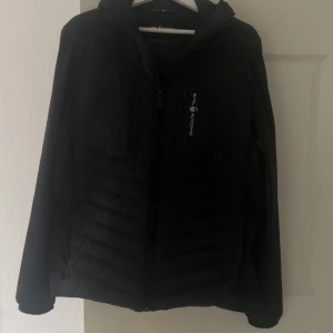 Svart dunjacka från Sail Racing - Svart dunjacka från Sail Racing med huva och snygga detaljer. Jackan har 800 fill power och är fodrad med Allied Feather + Down. Utrustad med dragkedja framtill och två bröstfickor med zip. Perfekt för kalla dagar och har en stilren look.