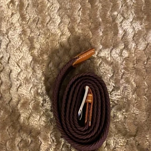 Mörkbrunt flätat bälte  - Snyggt mörkbrunt flätat bälte med detaljer i brunt skinn och silverspänne. Bältet har en stilren look och passar perfekt till jeans eller chinos. Flätad design gör det enkelt att justera längden efter behov.