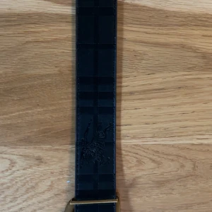 Burberry rutigt bälte med B-spänne - Säljer ett snyggt Burberry-bälte med klassiskt rutigt mönster. Ena sidan är svart med diskret rutig struktur och präglad ryttare, andra sidan är beige med bruna och svarta rutor samt guldig B-logga som spänne. Perfekt accessoar för att lyfta din outfit. Storleken är 115cm