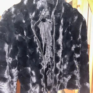 Svart pälsjacka från Soul River XS - Säljer en svart pälsjacka från Soul River i storlek XS. Jackan har en fluffig och mjuk yta med glansig finish och stängs med  framtill. Perfekt för dig som gillar att sticka ut med en cool och lyxig vibe. Fodrad insida för extra komfort.