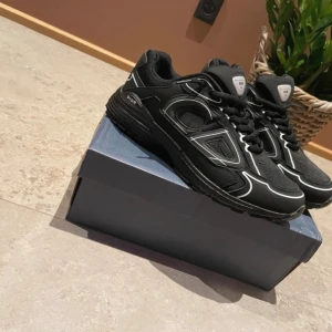 (Hjälper en vän) Svarta Dior B30 sneakers - Säljer ett par svarta Dior B30 sneakers med coola vita detaljer och CD-logga på hälen. Skorna har snörning, meshpaneler och en chunky platt sula. Märket syns tydligt på tungan och insidan. Perfekta för dig som gillar exklusiva och trendiga sneakers.