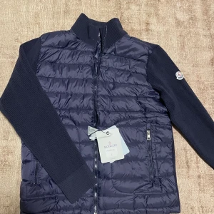 Mörkblå Moncler cardigan  - Snygg mörkblå pufferjacka från Moncler med quiltad front och stickade ärmar. Jackan har dragkedja, högt krage och Moncler-logga på ärmen. Insidan har coolt serietryck och Moncler-märke. Perfekt för dig som vill ha en stilren och exklusiv look. Den är i stl L men passar mer som M. Tags och allt medföljer. 