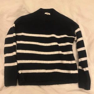 Randig svartvit stickad tröja - Snygg stickad tröja med svart botten och vita horisontella ränder. Tröjan har långärmad design och ribbad hals, ärmslut och nederkant. Perfekt för dig som gillar klassisk stil med en modern twist. Insydd vid armarna och sidorna på tröjan