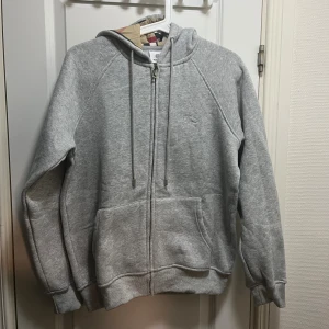 Grå hoodie med Burberry-mönstrad huva - Snygg grå hoodie från Burberry med klassiskt rutmönster i huvan. Tröjan har dragkedja, snörning och två stora fickor framtill. Tillverkad i mjuk bomull och har ribbade muddar vid ärmslut och nederkant. Perfekt för chill dagar.