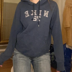 Vintage Nike hoodie med tryck - Snygg mörkblå hoodie från Nike med stort tryck 'NIKE 72' i grått på bröstet. Tröjan har huva med snörning, en klassisk känguruficka och långa ärmar. Den har några små fläckar på fickan men som man ser är de inget man märker
