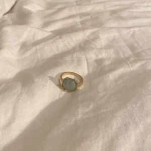 Guldfärgad ring med ljusblå sten - Snygg guldfärgad ring med en stor, ljusblå sten i sexkantig form. Ringen har en klassisk design med en unik touch tack vare den iögonfallande stenen som sitter i en elegant fattning. Perfekt för dig som vill sticka ut med ett statement-smycke.
