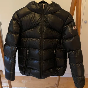 Svart Colmar pufferjacka med huva - Svart pufferjacka från Colmar med glansig yta och huva. Köpt för 6999 kr. Jackan har två sidofickor med dragkedja, markerade sömmar och Colmar-logga på ärmen. Perfekt för kalla dagar och har en snygg, modern look med lång ärm och rak passform. Pris kan diskuteras. Hör av er vid frågor! 🥰🥰
