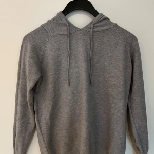 Grå stickad hoodie med huva - Mysig grå stickad hoodie från Blend med dragsko i huvan och långa ärmar. Tröjan har en enkel och clean look, perfekt för chill dagar. Ribbstickade muddar vid ärmslut och nederkant ger en snygg passform.