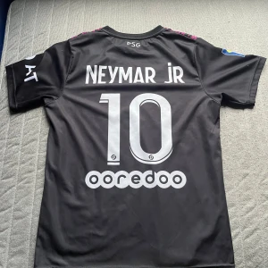 PSG Neymar Jr fotbollströja Nike - Säljer en PSG fotbollströja med Neymar Jr och nummer 10 på ryggen. Tröjan är svart med korta ärmar och har ett unikt mönster framtill i rosa och svart med en stor orm över bröstet. Märkt med Qatar Airways och PSG-logga. Tillverkad i lätt och ventilerande material.