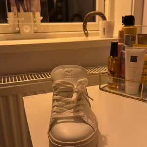 Vita sneakers från Veja strl 38 - Snygga vita sneakers från Veja i storlek 38. Skorna har klassisk rund tå, platt sula och snörning framtill. Tillverkade i skinn med beige detaljer på sidan och hälen. Perfekt för dig som gillar stilrena och trendiga sneakers med minimalistisk design.
