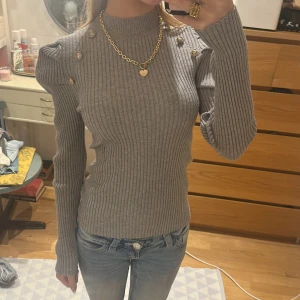 Grå ribbad tröja från Emery Rose - Supersnygg grå ribbad tröja från Emery Rose med markerade axlar och dekorativa guldknappar på axlarna. Tröjan har lång ärm, hög hals och figurnära passform. Mindre i storlek men är stretchig så passar även S!!🫶
