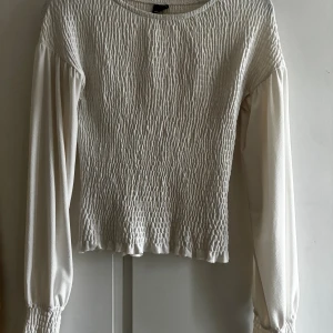 Vit blus med smock från Gina Tricot - Supersöt vit blus från Gina Tricot i storlek XS. Toppen har smockad kropp och långa puffärmar med resår vid ärmslut. Rund halsringning och lätt volangkant nedtill ger en extra touch. Perfekt för dig som gillar romantisk och trendig stil.