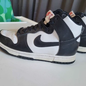 Panda Nike Dunk High svart/vit - Nike Dunk High sneakers i svart och vitt skinn med klassisk svart swoosh på sidorna. Skorna har snörning, perforerad tå och hög siluett. Yttersulan är svart och vit, och plösen har en röd Nike-logga. Perfekta för dig som gillar retrostil.
