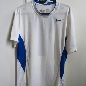 Nike Pro Combat vit/blå tränings-tshirt - Nike Pro Combat Dri-Fit t-shirt i vitt med blåa meshpaneler på sidorna och ryggen. Kortärmad och med rund hals, designad för träning med ventilerande material och fitted passform. Snyggt blått Nike-logga på bröstet och coolt mönster på ryggen.Tröjan är ny utan prislapp.