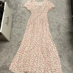 Blommig omlottklänning med volang - Säljer en lång, vit omlottklänning med röda och rosa blommor. Klänningen har korta puffärmar, v-ringning och volangdetaljer längs kjolen. Perfekt för sommarens alla tillfällen och har en romantisk vibe. Materialet känns lätt och luftigt.