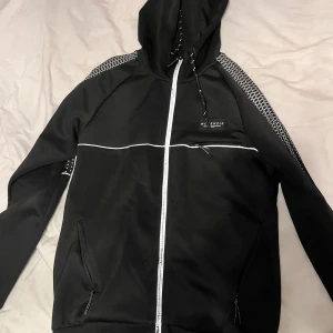 Svart vindjacka från McKenzie - Svart vindjacka med huva från McKenzie. Jackan har vita detaljer, dragkedja framtill och snörning i huvan. Snyggt mönstrade axelpartier och två fickor med dragkedja. Perfekt för en sportig stil. Lite trasig i tråden se bild