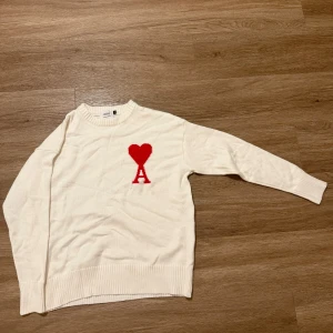 Vit stickad tröja från Ami - Vit stickad tröja från Ami Alexandre Mattiussi med rund halsringning och ribbade muddar. På bröstet finns ett rött hjärta med bokstaven A. Perfekt för dig som gillar stilrena plagg med en twist.