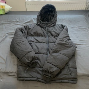 Svart pufferjacka med huva - Svart pufferjacka med huva och snörning, perfekt för kalla dagar. Jackan har långa ärmar med resår vid mudden, två sidofickor med dragkedja och quiltad design. Materialet är syntet och jackan stängs med dragkedja framtill.