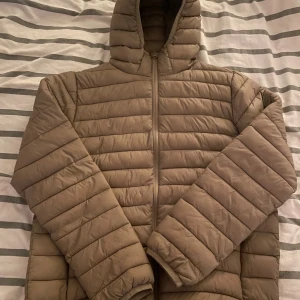 Beige pufferjacka från H&M Divided - Säljer en beige pufferjacka från H&M Divided i storlek L. Jackan har huva, horisontella quiltade paneler och dragkedja framtill. Perfekt för kyliga dagar och har en clean, modern look som funkar till många stilar.