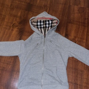 Grå hoodie från Burberry med dragkedja - Grå hoodie från Burberry med klassiskt rutigt foder i huvan och broderad logga på bröstet. Tröjan har dragkedja framtill, snörning vid huvan och två fickor. Perfekt för dig som gillar stilren design med en twist.